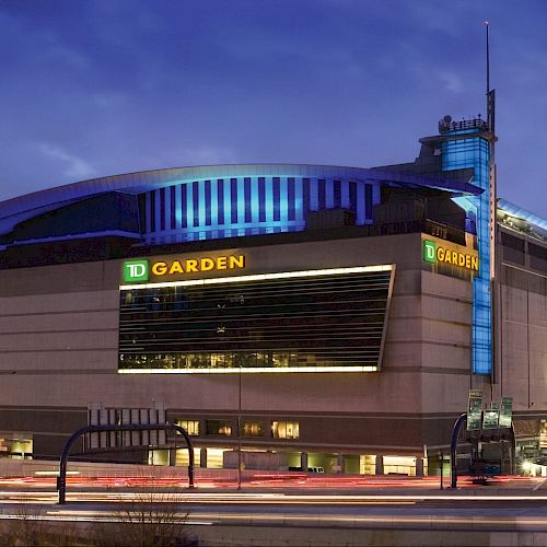 The image shows an exterior view of TD Garden, an arena in a city setting with the skyline illuminated against a twilight sky.