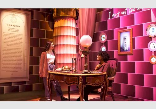 Two people are interacting in a themed room with a vintage desk, a lamp, a picture, and decorative plates on pink walls ending the sentence.