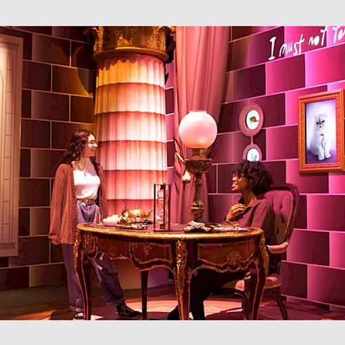 Two people are interacting in a themed room with a vintage desk, a lamp, a picture, and decorative plates on pink walls ending the sentence.