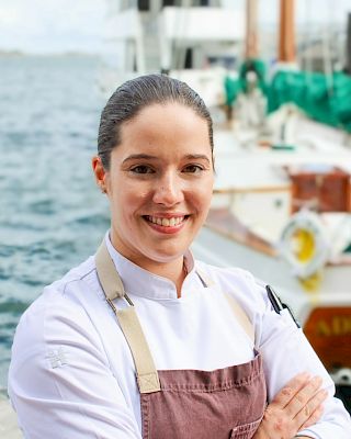 A person in a chef's uniform stands by the waterfront with boats in the background, smiling at the camera.