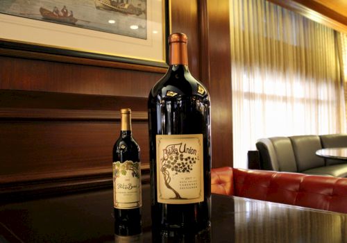 The image shows two wine bottles, one much larger than the other, on a polished table in a room with wood paneling and a framed picture.