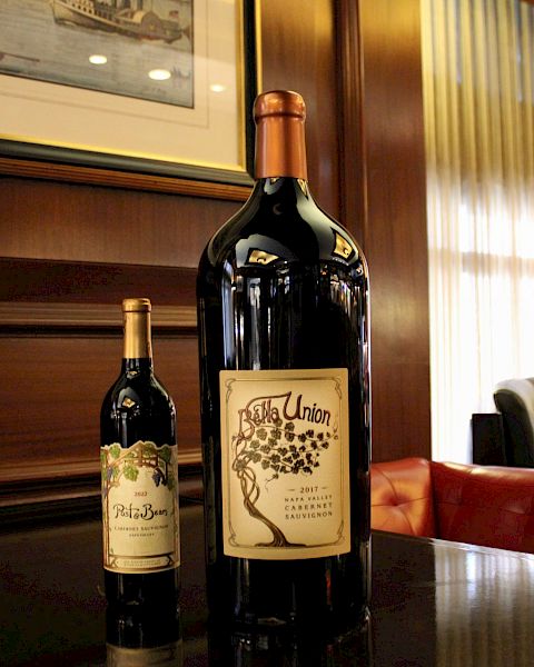 The image shows two wine bottles of different sizes on a polished table in a wood-paneled room with a sofa and art on the wall.