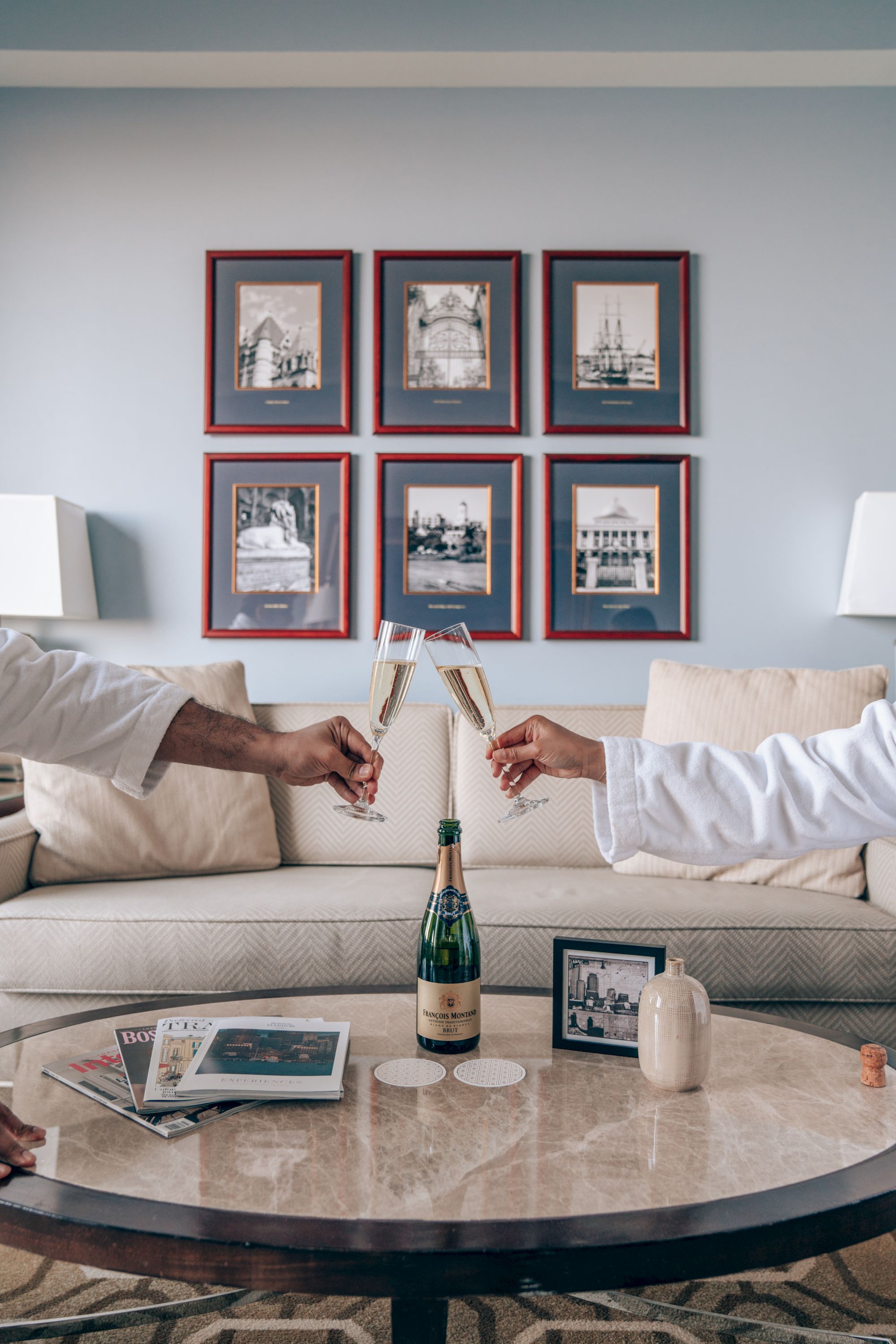 Two people in robes toast with champagne flutes over a table with a champagne bottle, books, and a framed photo in a living room.