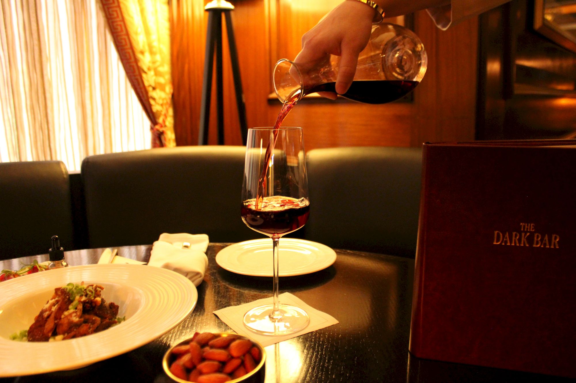 A hand pours red wine into a glass on a table set with food, almonds, and a menu labeled 