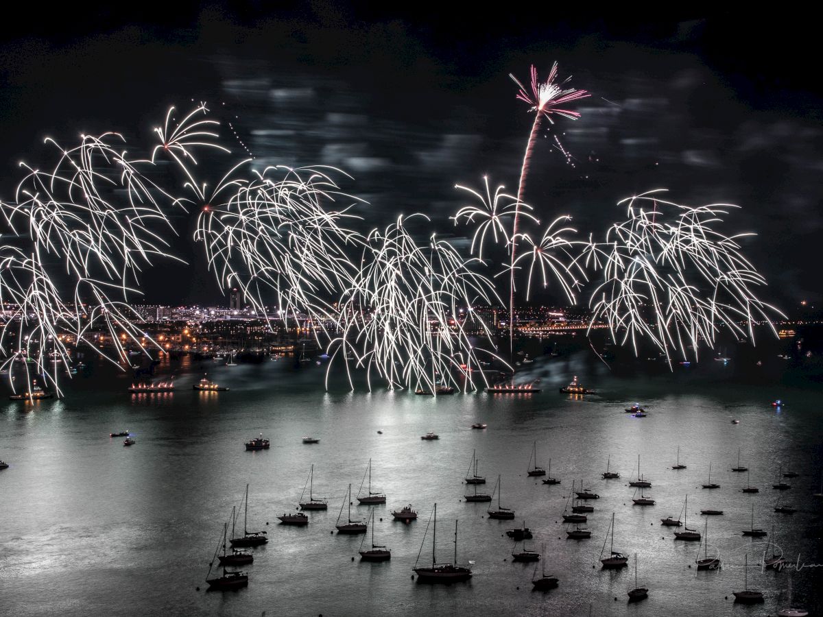 Fireworks light up the night sky over a harbor, with numerous boats anchored in the water reflecting the vibrant display.