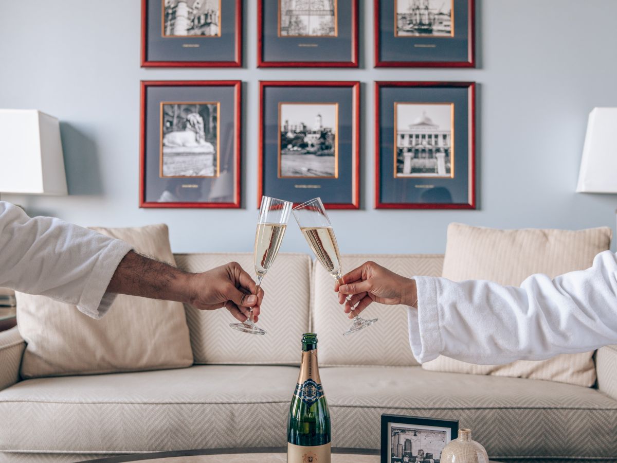 Two hands in robes clink champagne glasses over a table with books and a bottle, set in a cozy room with framed pictures on the wall.