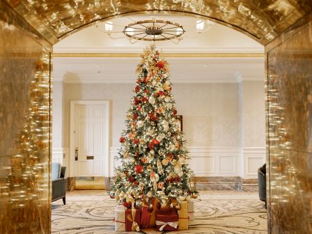 A decorated Christmas tree stands in an elegant room, adorned with lights, ornaments, and surrounded by gifts.