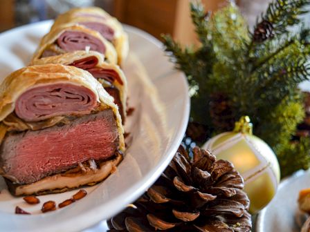 The image shows slices of beef Wellington on a plate, garnished with pinecones and a Christmas ornament, creating a festive setting.