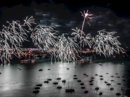 Fireworks illuminate the night sky over a calm bay dotted with boats, reflecting on the water, creating a festive and vibrant scene.