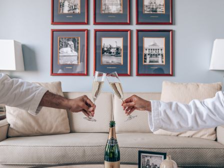 Two people in robes toast with champagne flutes over a table with books and a bottle, in a room with framed pictures on the wall.