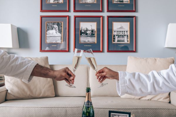 Two people in robes toast with champagne flutes over a table with books and a bottle, in a room with framed pictures on the wall.