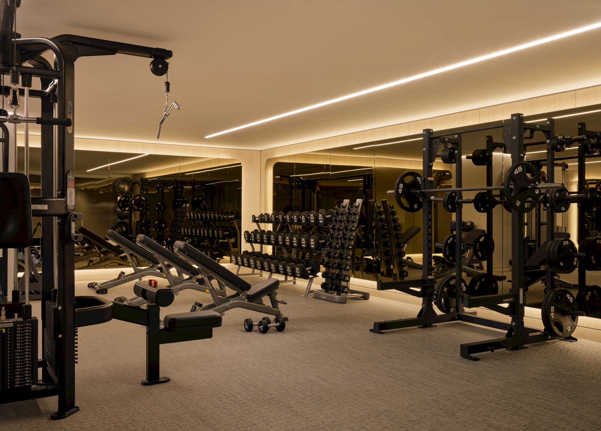 The image shows a well-lit gym with weightlifting equipment, benches, and mirrors lining the walls.