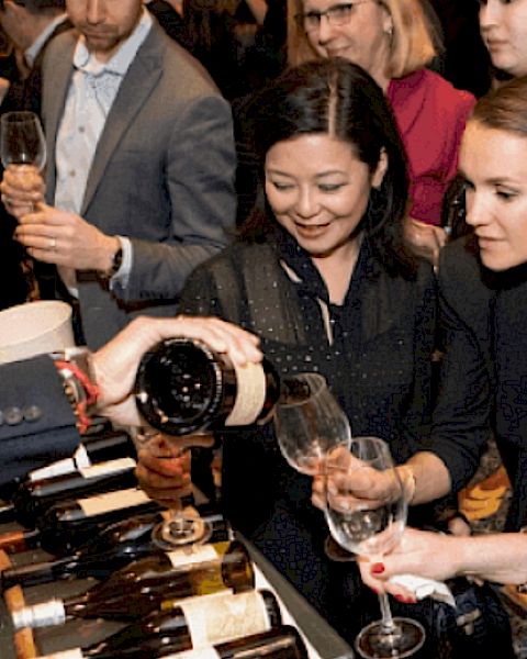 People are gathered at an event, tasting wine as a server pours from a bottle into their glasses.