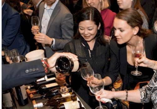 People are gathered at an event, tasting wine as a server pours from a bottle into their glasses.