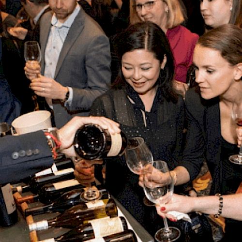 People are gathered at an event, tasting wine as a server pours from a bottle into their glasses.