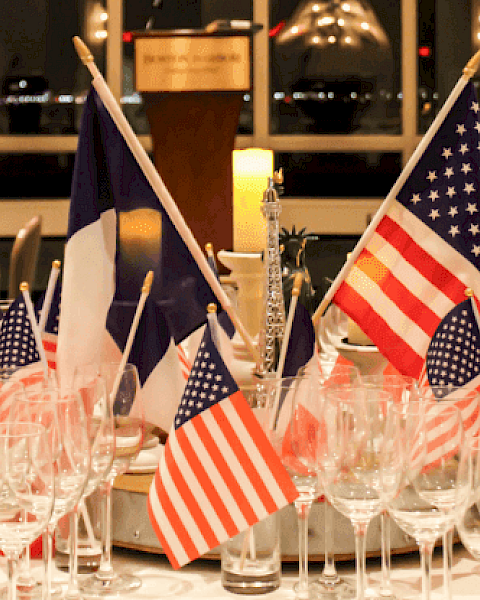 The image shows a table centerpiece with American and French flags, surrounded by empty glasses, set in a formal dining setting.