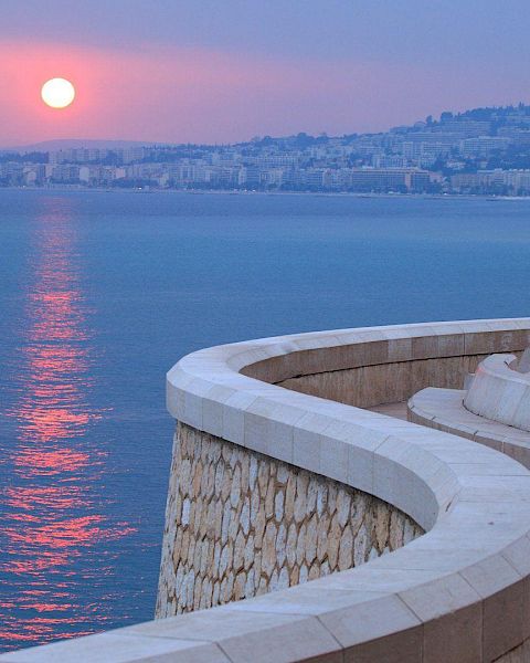 A winding stone pathway curves along a waterfront, with a city skyline and setting sun reflected on the calm sea, under a purple-toned sky.