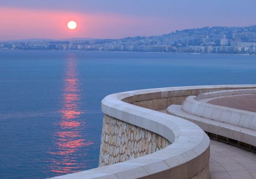 A winding stone pathway curves along a waterfront, with a city skyline and setting sun reflected on the calm sea, under a purple-toned sky.