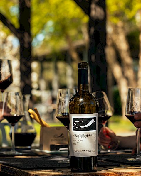 A group of people enjoying wine outdoors around a table, with a bottle of wine prominently displayed in the center.