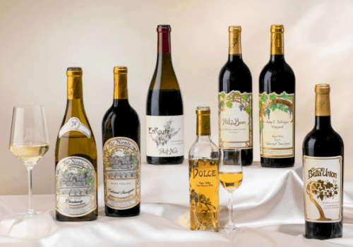 A selection of seven wine bottles with varying labels, alongside two filled wine glasses, set on a white cloth background.