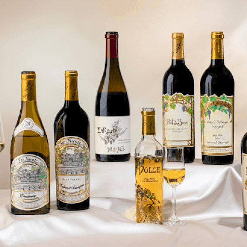 A selection of seven wine bottles with varying labels, alongside two filled wine glasses, set on a white cloth background.