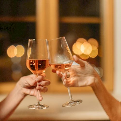 Two people clinking glasses of rosé wine in a warmly lit setting, with blurred lights in the background creating a festive atmosphere.