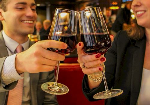 Two people clinking wine glasses in a celebratory manner, sitting in a cozy setting with smiles and dressed in formal attire.