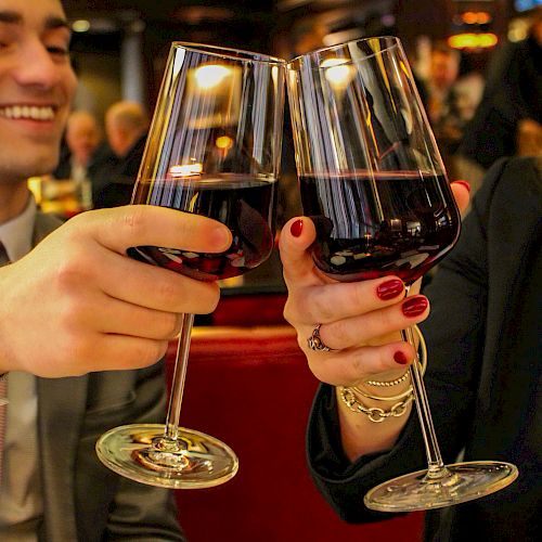 Two people clinking wine glasses in a celebratory manner, sitting in a cozy setting with smiles and dressed in formal attire.