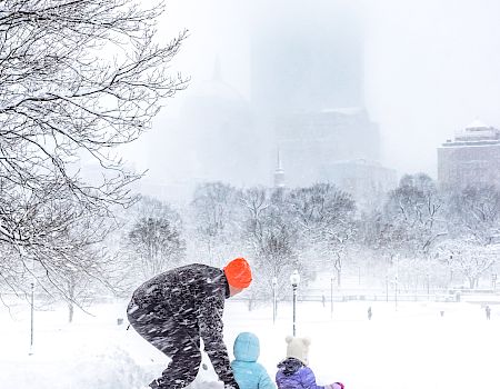 A person and two children play in the snow during a heavy snowfall, with blurry city buildings visible in the background.