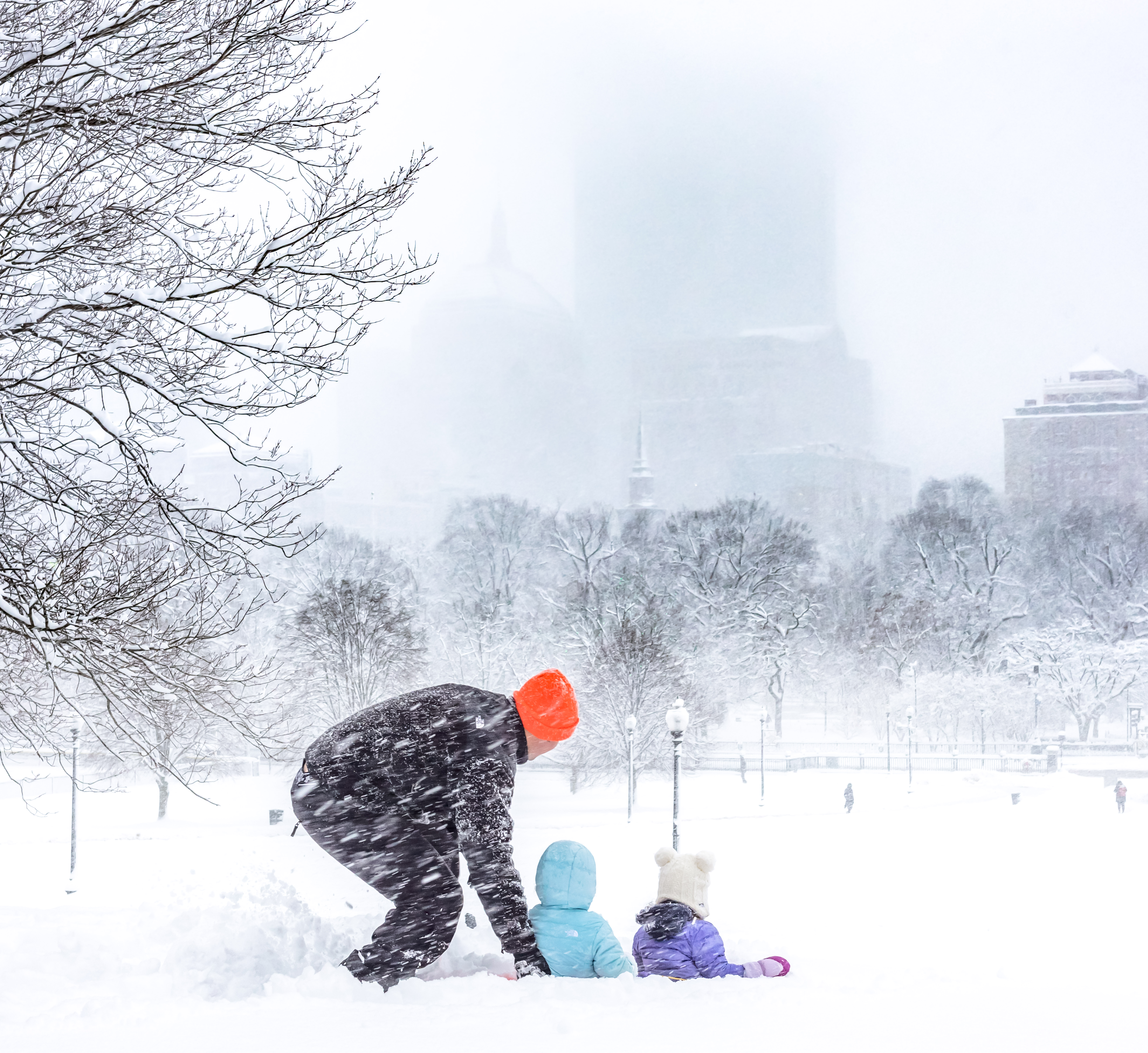 A person and two children play in the snow during a heavy snowfall, with blurry city buildings visible in the background.