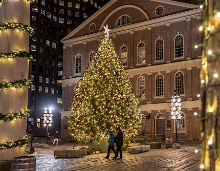 A large Christmas tree adorned with lights and ornaments stands in front of a historic building, surrounded by festive decorations.