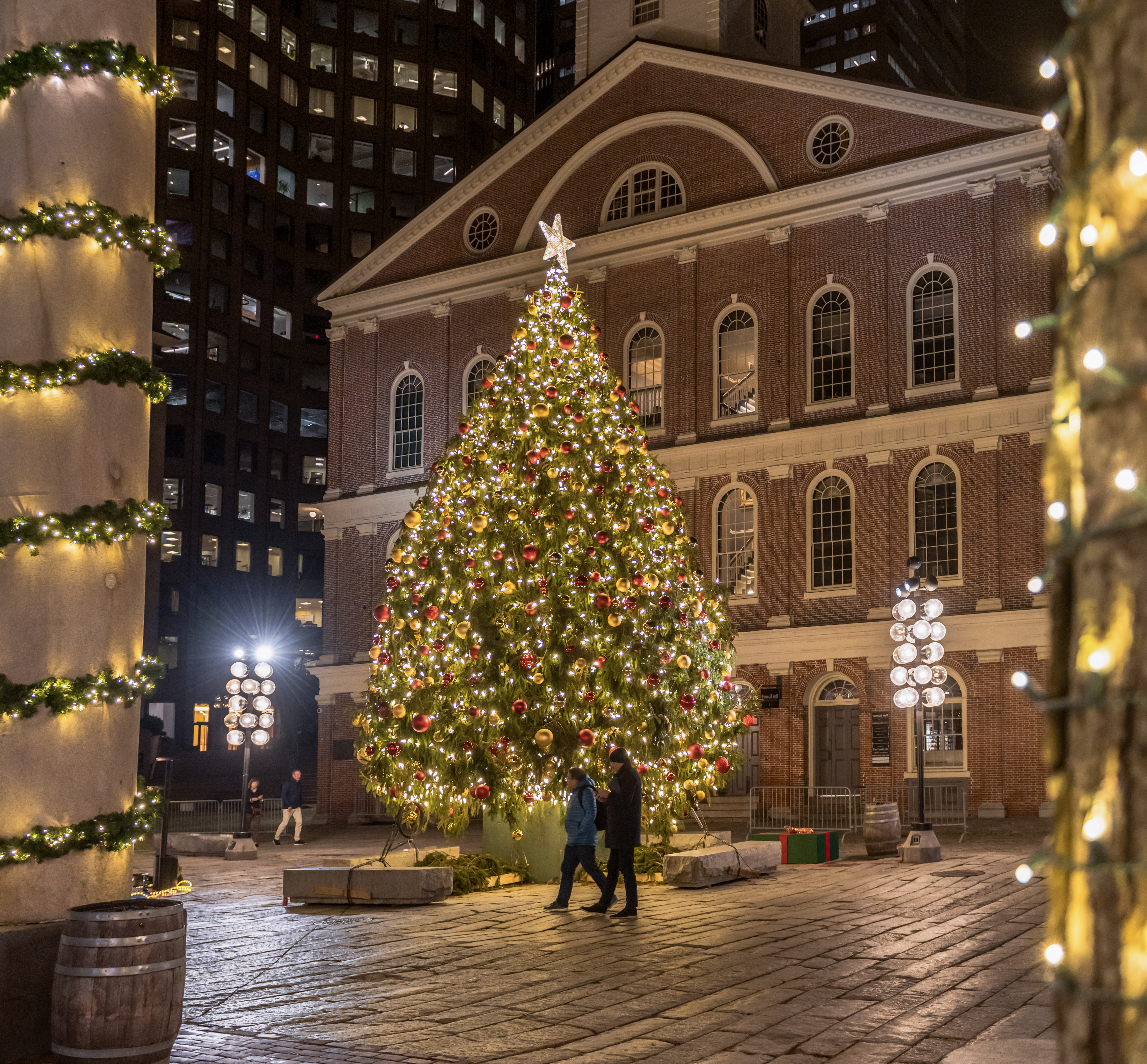 A large Christmas tree adorned with lights and ornaments stands in front of a historic building, surrounded by festive decorations.