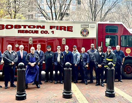 A group of firefighters stands in front of a Boston Fire Rescue Co. 1 truck on a city street with trees and buildings in the background.