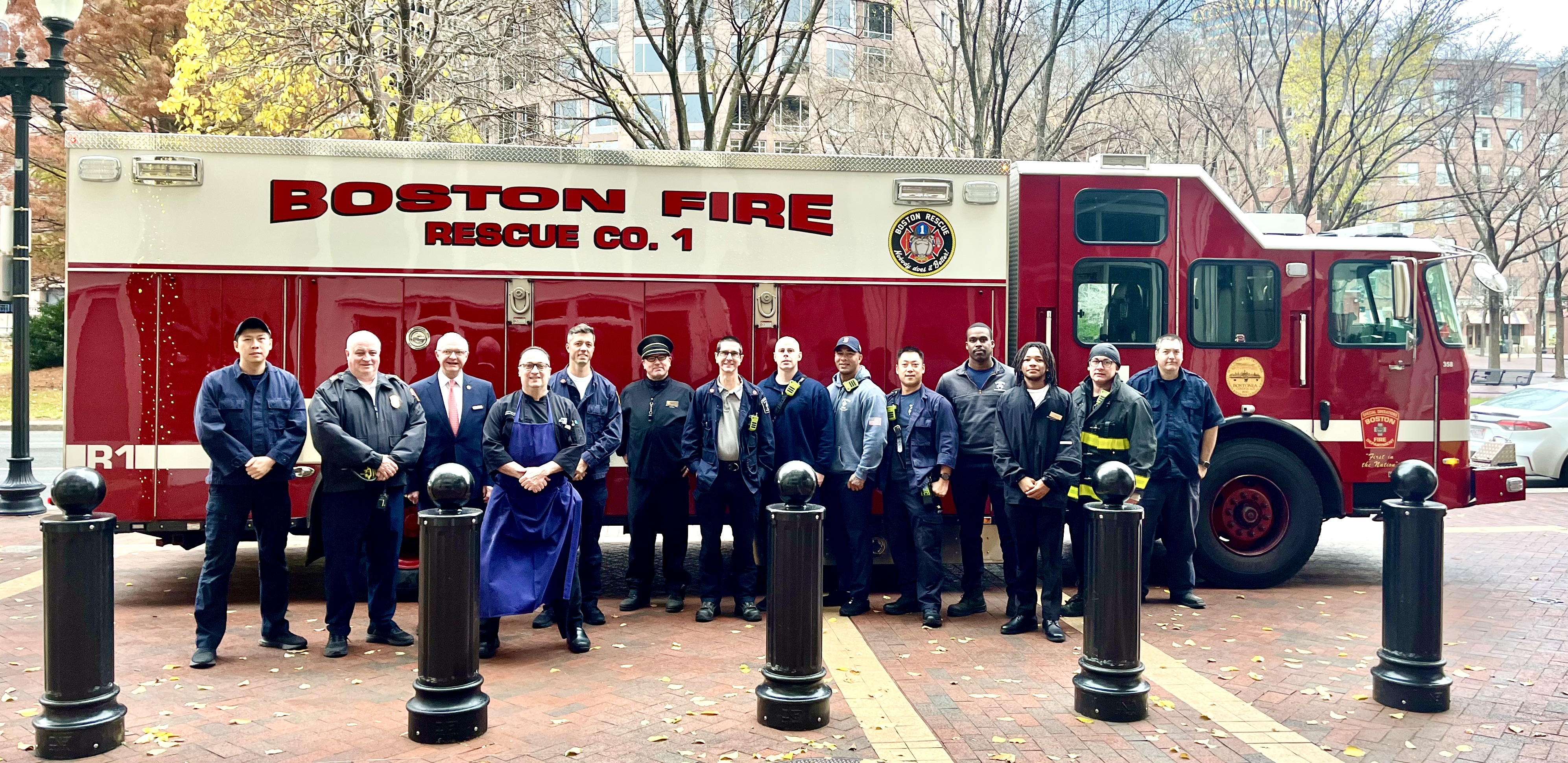 A group of firefighters stands in front of a Boston Fire Rescue Co. 1 truck on a city street with trees and buildings in the background.