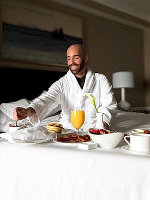 A person in a bathrobe enjoys breakfast in bed with orange juice, fruits, and pastries on a tray in a hotel room.