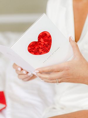 A person in white holds a card with a red heart. An envelope lies nearby on a bed.