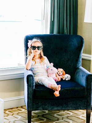 A child with sunglasses sits on a blue chair, holding stuffed animals, in a room with patterned carpet and a large window.