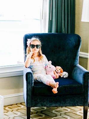 A child with sunglasses sits on a blue chair, holding stuffed animals, in a room with patterned carpet and a large window.