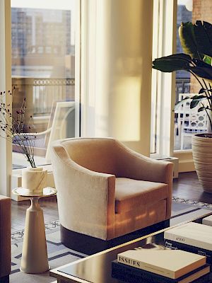 A modern living room with beige chairs, a large potted plant, a coffee table with books, and floor-to-ceiling windows.