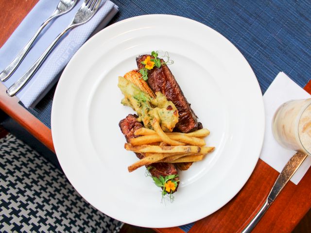 The image shows a plated dish with steak, fries, garnished with edible flowers, on a table setting with cutlery and a drink.