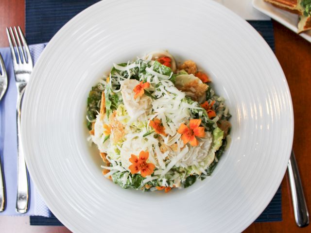 A salad with shredded cheese, greens, and orange edible flowers on a white plate. Forks and a small dish are visible nearby.