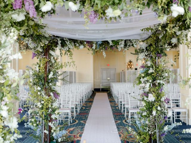 A floral archway leads to an aisle lined with white chairs in a beautifully decorated indoor wedding venue ending the sentence.