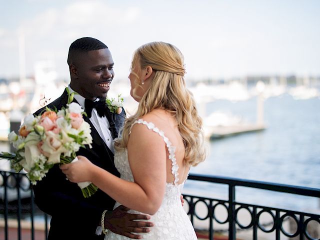 A couple in wedding attire smiles at each other near a waterfront, holding a bouquet.