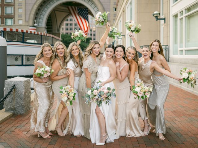 A group of women in formal dresses holding bouquets, smiling and posing joyfully outdoors with a large archway and flag in the background.