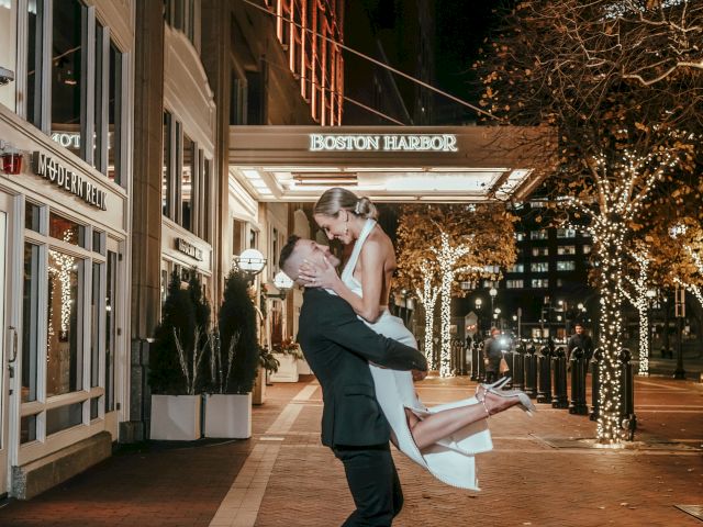 A couple is outside at night, with the man lifting the woman high in a joyful pose outside a well-lit building adorned with trees and lights.