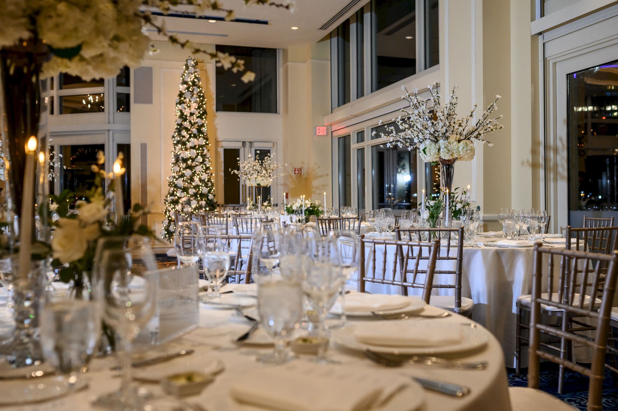 A beautifully decorated banquet hall with elegantly set tables, floral arrangements, and a lit tree, creating a festive ambiance.