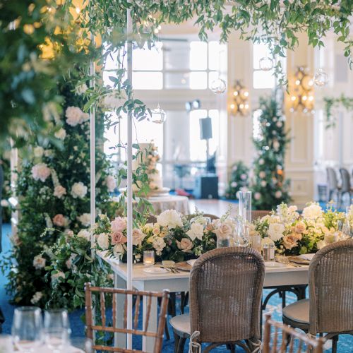 A beautifully decorated indoor event space with elegant floral arrangements, hanging greenery, and tables set with dishes and glassware.