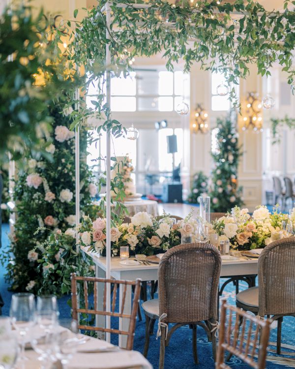 A beautifully decorated indoor event space with elegant floral arrangements, hanging greenery, and tables set with dishes and glassware.