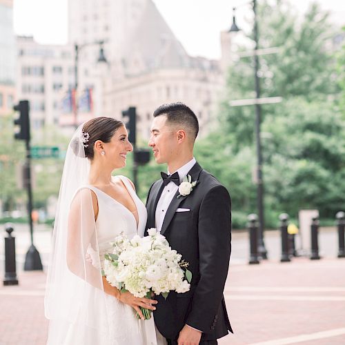 A bride in a white dress and a groom in a suit stand joyfully with a cityscape backdrop, holding a bouquet of white flowers.