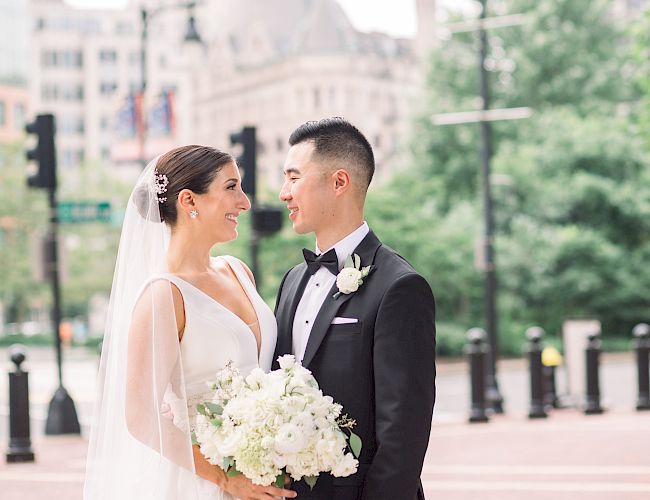 A bride in a white dress and a groom in a suit stand joyfully with a cityscape backdrop, holding a bouquet of white flowers.